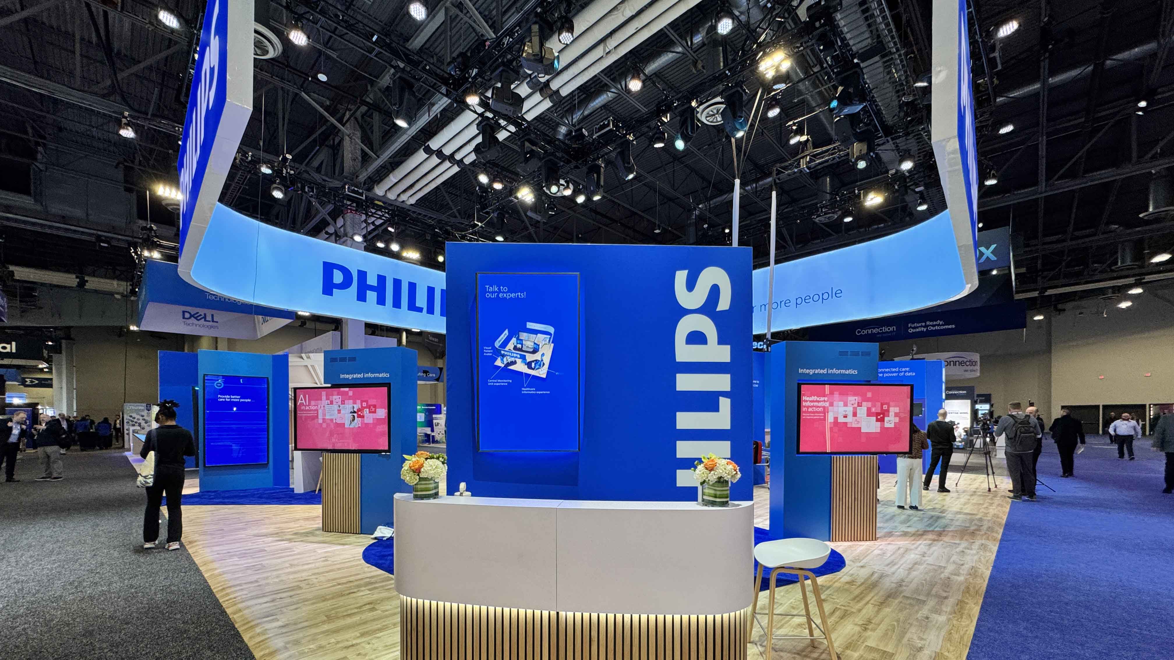 An image of a tradeshow booth. The booth is blue in color and has a large screen at the center. The booth is Owned by Philips
