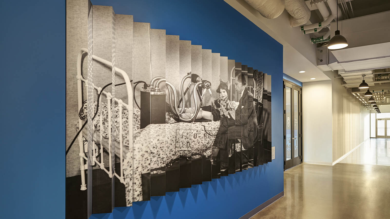 A Photograph of a lenticular image on a wall in a hall way. The lenticular image has a black and white image of a historical ultrasound maachine.