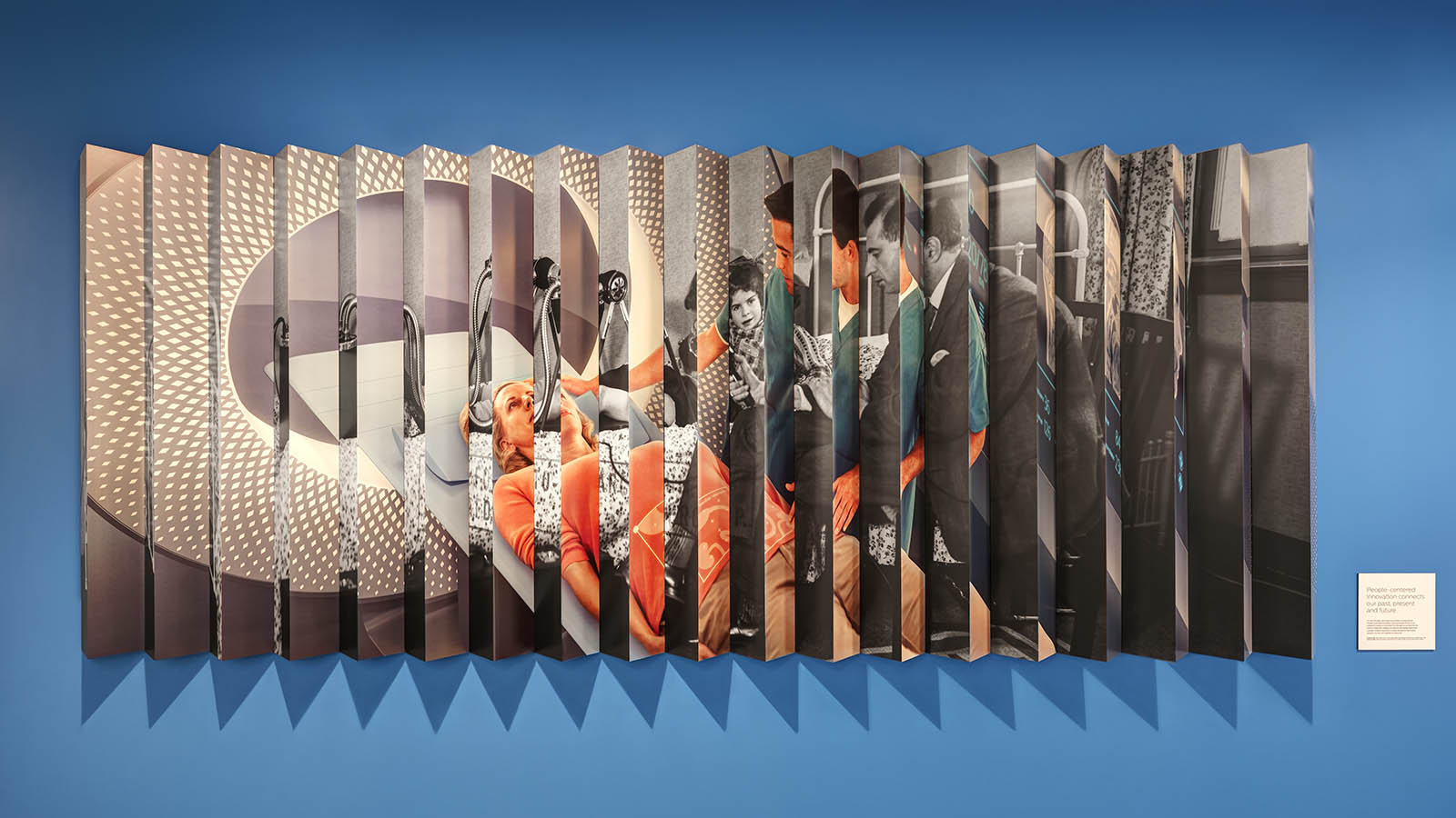 A Photograph of a lenticular image on a wall with a description in a small square on the right side. The lenticulare image has a black and white image of a historical ultrasound maachine and an image of a futuristic CT or MR machine in color.